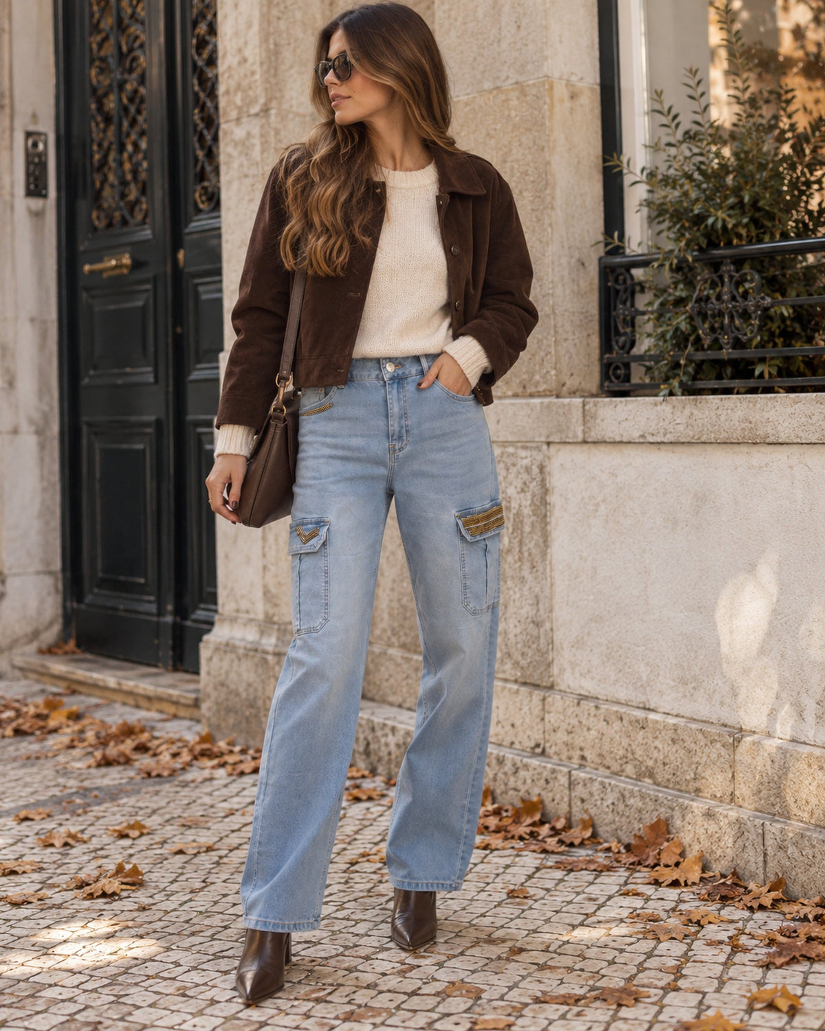 Calças cargo em ganga azul-clara com missangas douradas, em look de dia frio com malha creme, casaco castanho e botins
